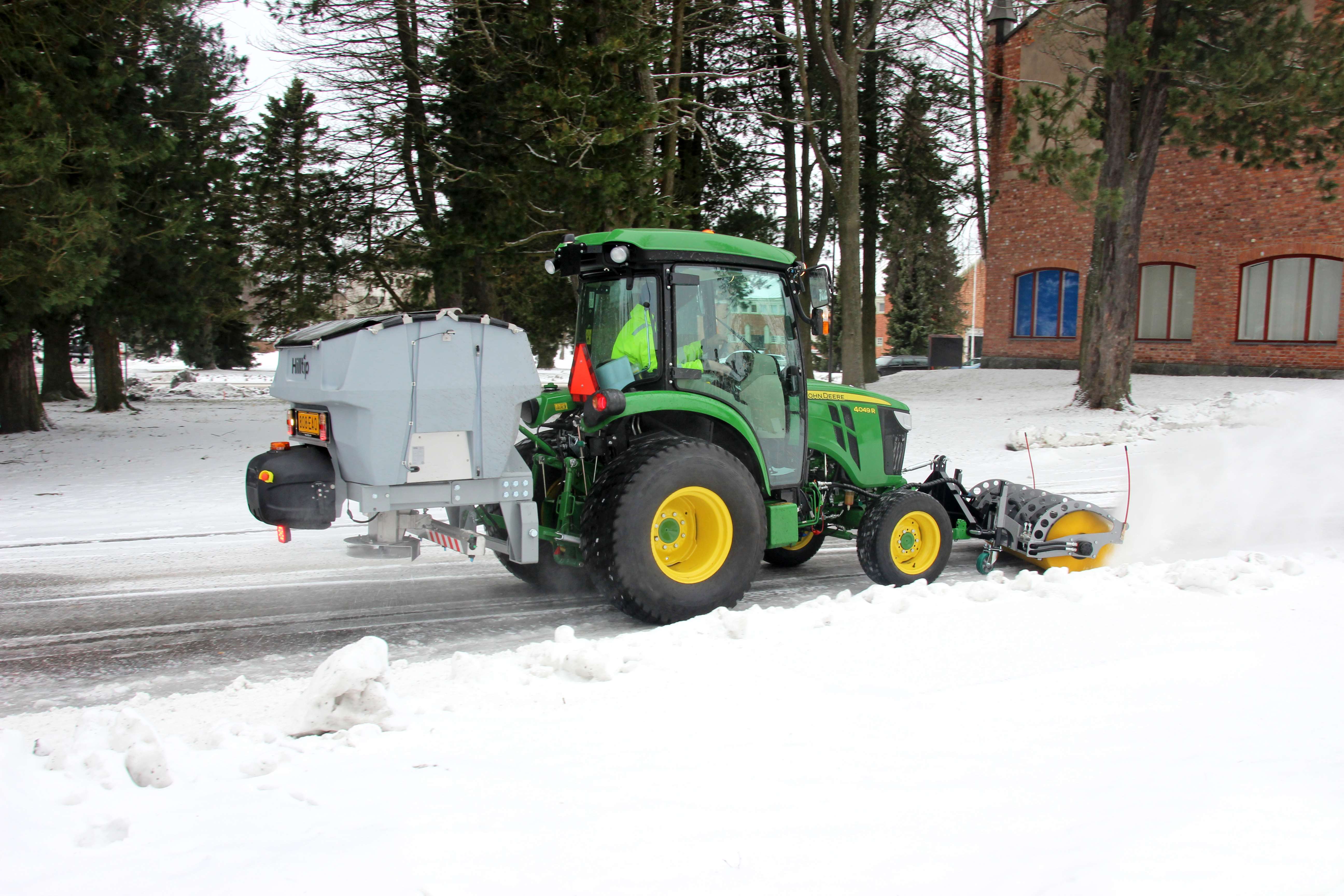 John Deere tractor equipped with rotary broom and salt spreader clearing snow on a winter road near trees and a brick building.